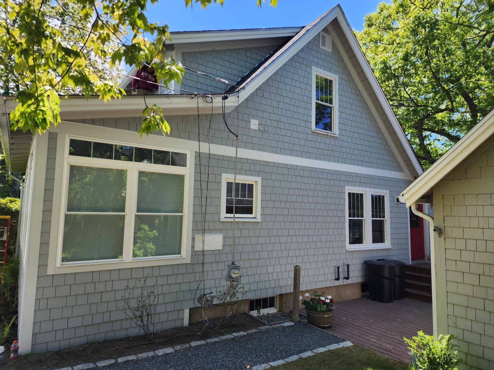 Blue house with a steep roof, several windows, black bins on the porch, and trees surrounding.