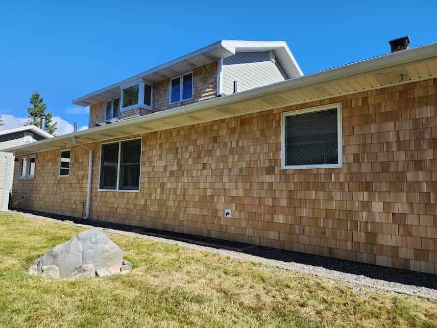 A long two-story home with cedar shingle siding, dark window, and a large rock in the yard.