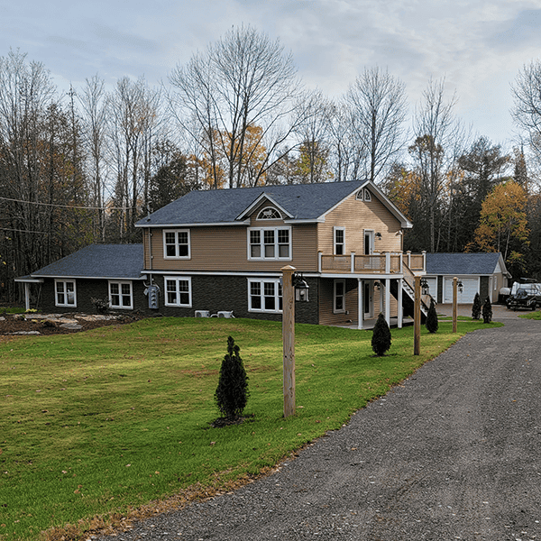 A two-story home with grey roof, brown vinyl siding, white-framed windows, a second-story deck, surrounded by trees, a large yard and driveway.