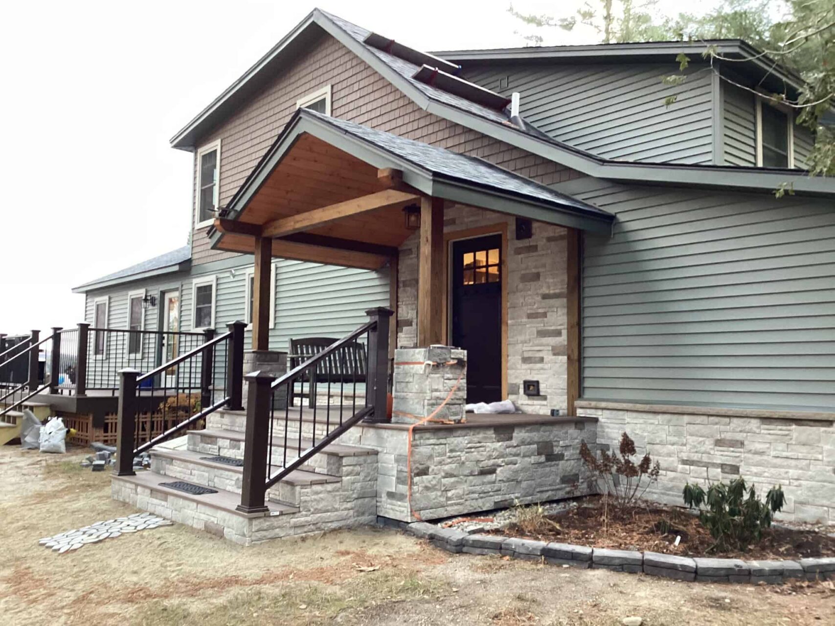 A two-story home with green vinyl siding, tan and grey stone veneer, and two decks that have black metal railings.