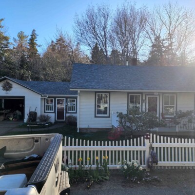 A white house with a garage, black roof, and windows, surrounded by green trees and a white picket fence. A truck in the drive.