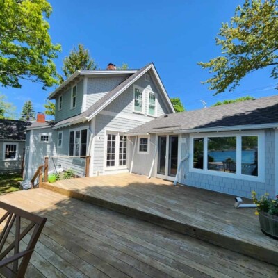 Light blue house with blue siding, a black asphalt roof, and large wooden deck. It has multiple windows, including a big one with white trim.