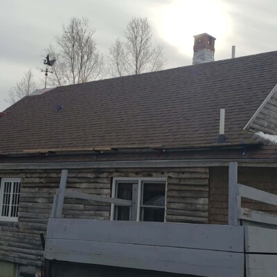 Wooden log cabin with a brown asphalt shingle roof, two windows, and a chimney, set against a backdrop of bare tree branches.